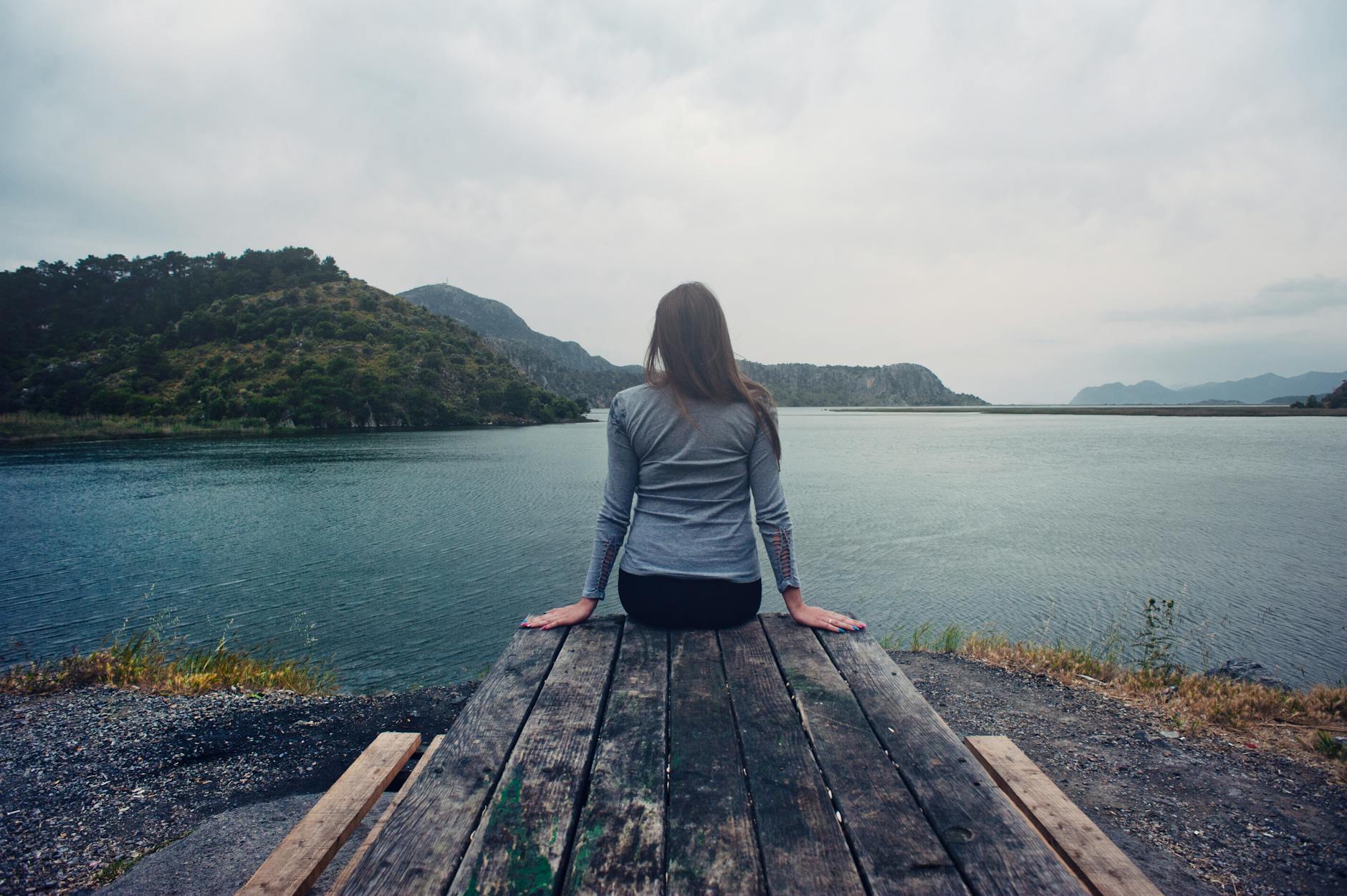 woman wearing gray long sleeved shirt and black black bottoms outfit sitting on gray wooden picnic table facing towards calm body of water at daytime